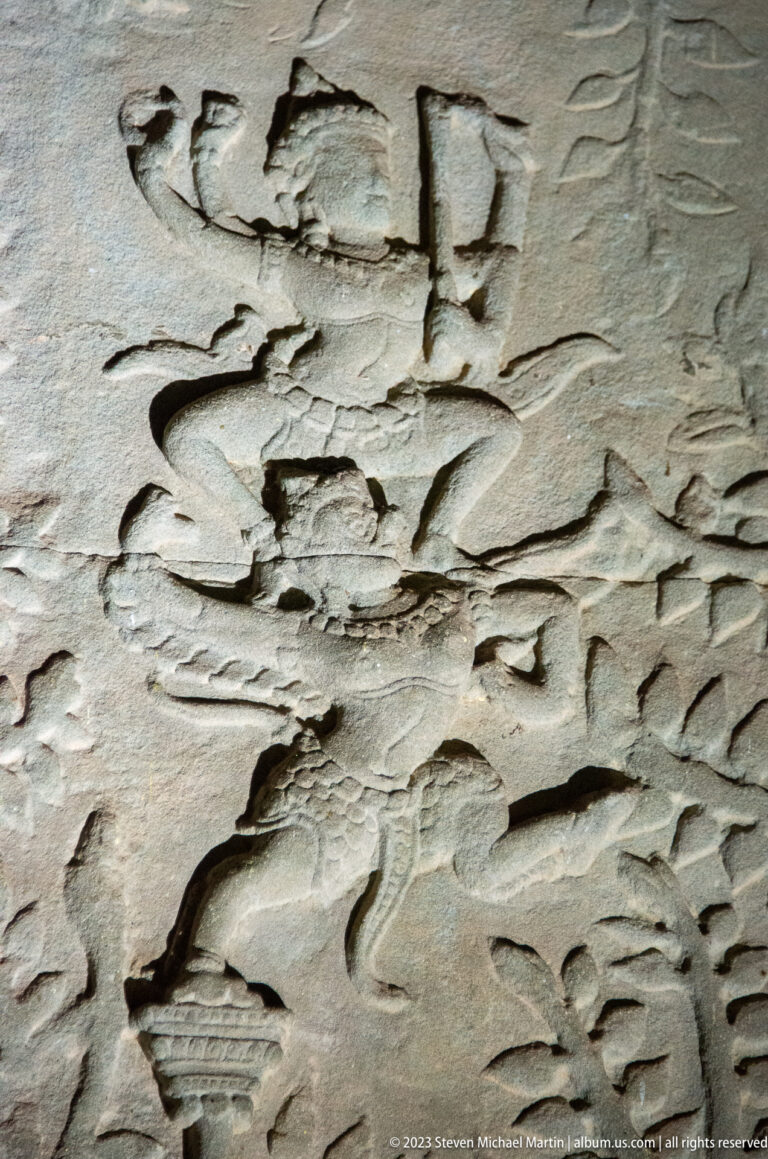Weathered relief showing a figure above a long carved vessel or platform at Angkor Wat.