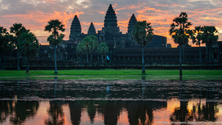 Closer dawn view of the five tower silhouette with palms and a bright orange horizon reflected in water at Angkor Wat.