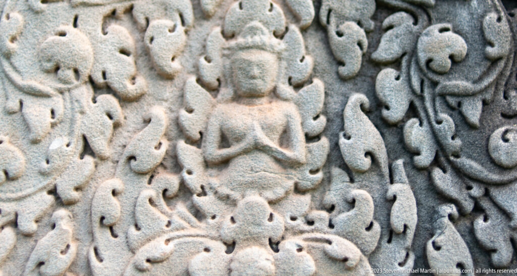 Small seated Buddha or sacred figure carved within a dense curling floral surround at Angkor Wat.