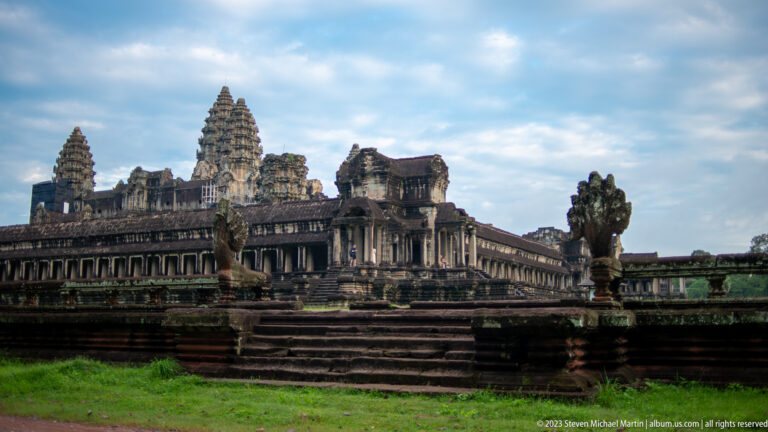 Wide oblique exterior of Angkor Wat with layered steps, long gallery, towers, and a guardian at right at Angkor Wat.