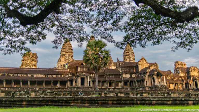Broad frontal façade framed by overhanging tree branches and late-afternoon light at Angkor Wat.