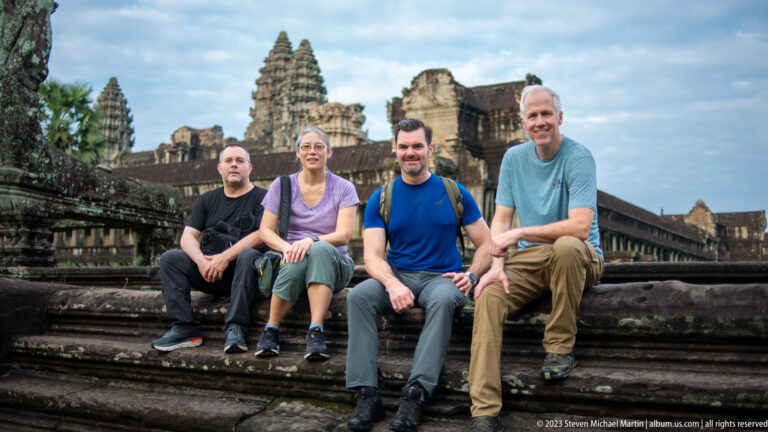 Four visitors seated together on broad stone steps with towers behind at Angkor Wat.
