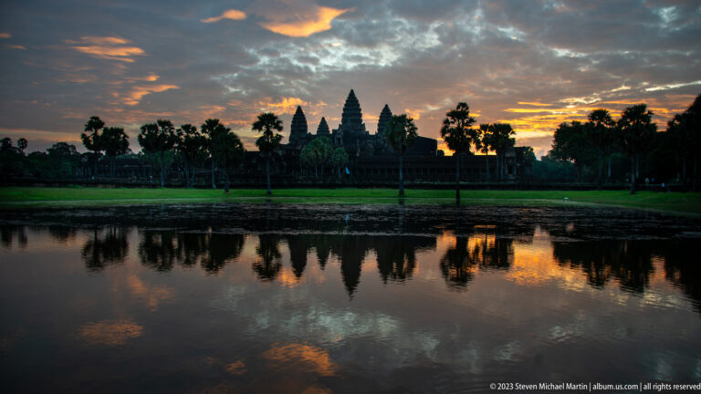 Dark Angkor Wat silhouette and palm trees mirrored in the reflecting pool beneath glowing clouds at Angkor Wat.