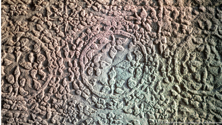 Dark weathered decorative panel with a circular central figure surrounded by dense repeated carving at Angkor Wat.