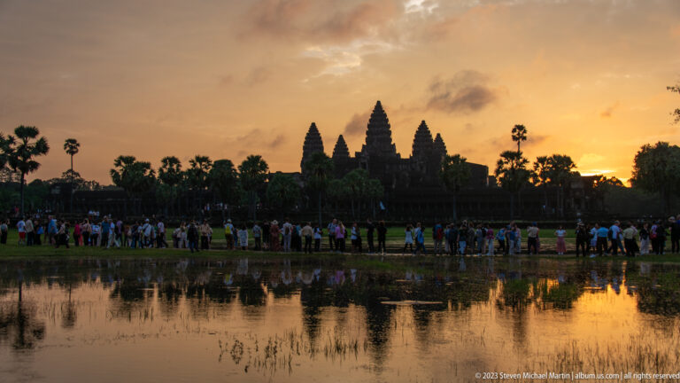 Angkor Wat silhouetted at sunrise with a crowd along the water’s edge and gold reflection at Angkor Wat.
