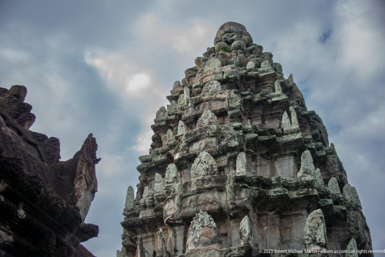 Weathered tiered sanctuary tower against cloudy blue sky at Angkor Wat.