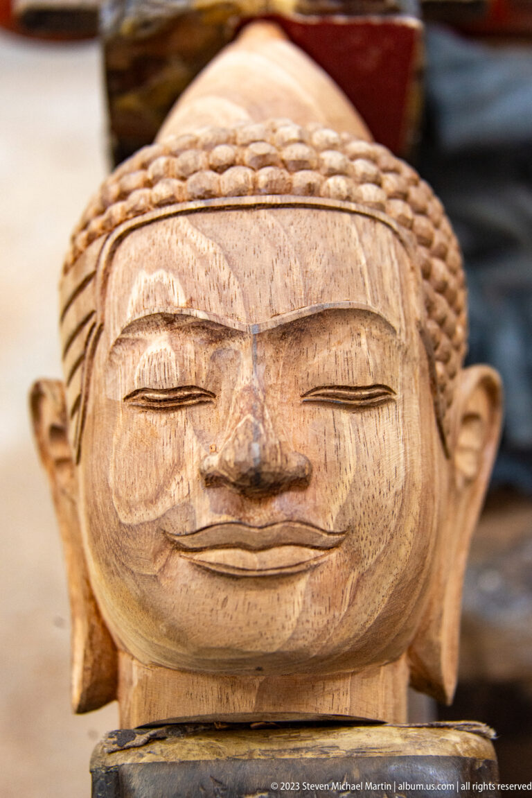 Close frontal view of an unfinished wooden Buddha face with closed eyes and visible grain.