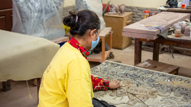 Masked woman in a yellow shirt rubbing the surface of a large decorated relief panel.