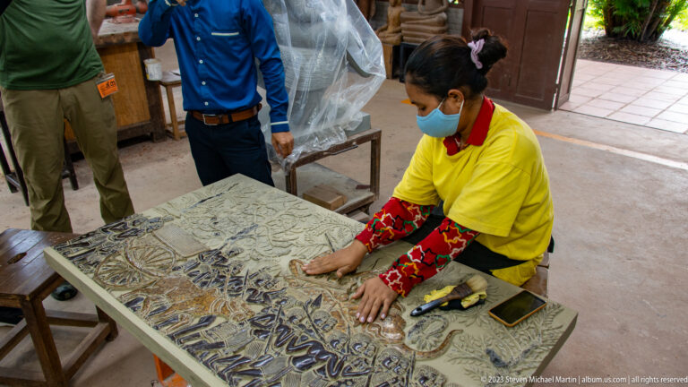 Masked artisan finishing a large relief panel while two visitors stand beside the worktable.