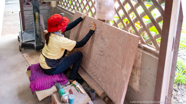 Red-hatted artisan tracing a design onto a large upright pink relief panel.
