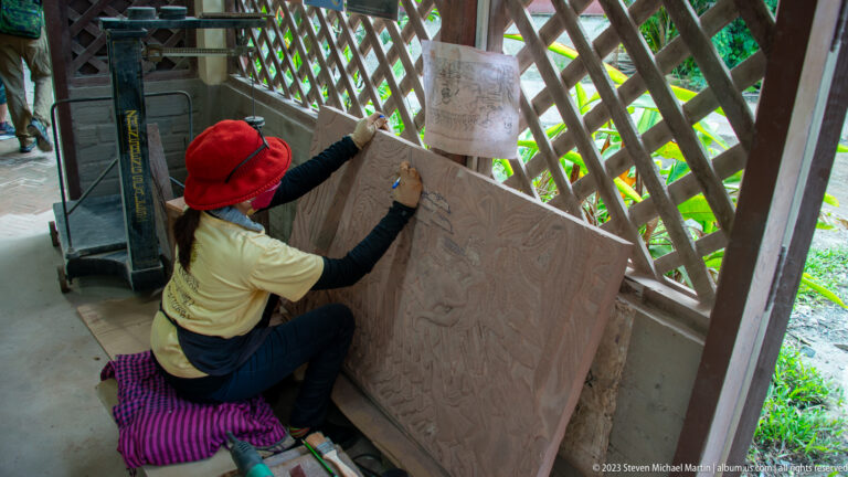 Side view of a red-hatted artisan reaching across a large incised relief panel.
