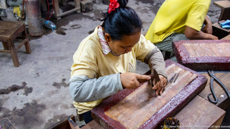 Woman hammering a punch into a patterned metal sheet on a thick reddish support block.