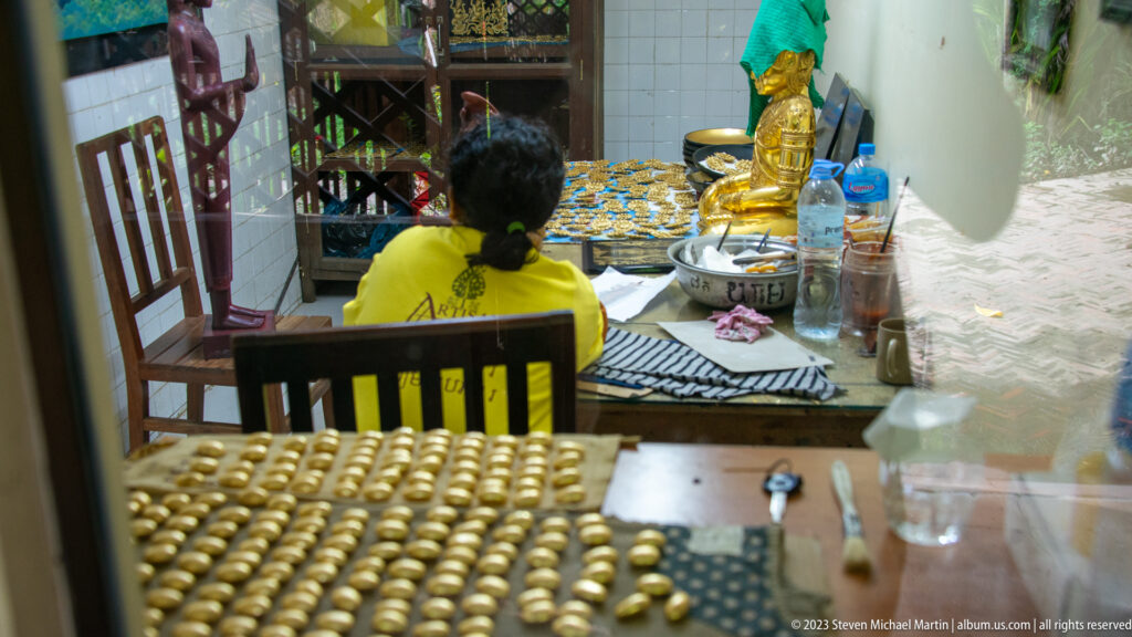 Rows of gold-colored oval pieces in front of an artisan working at a crowded table.