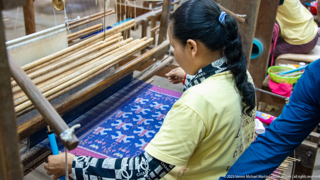 Woman weaving a vivid blue textile with red and white elephant motifs on a wooden loom.