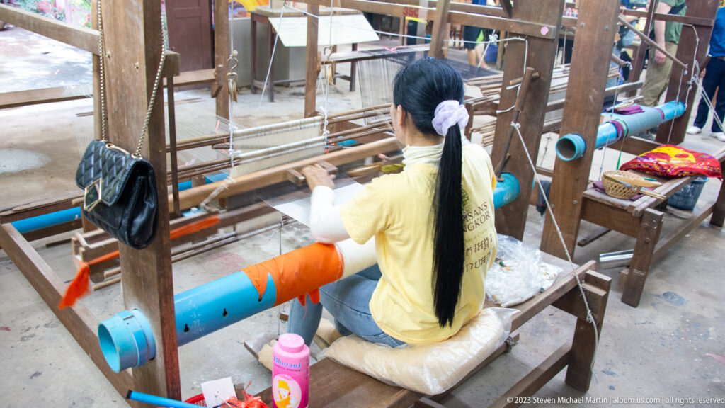 Rear view of a woman seated at a large wooden loom with orange and blue thread rollers.