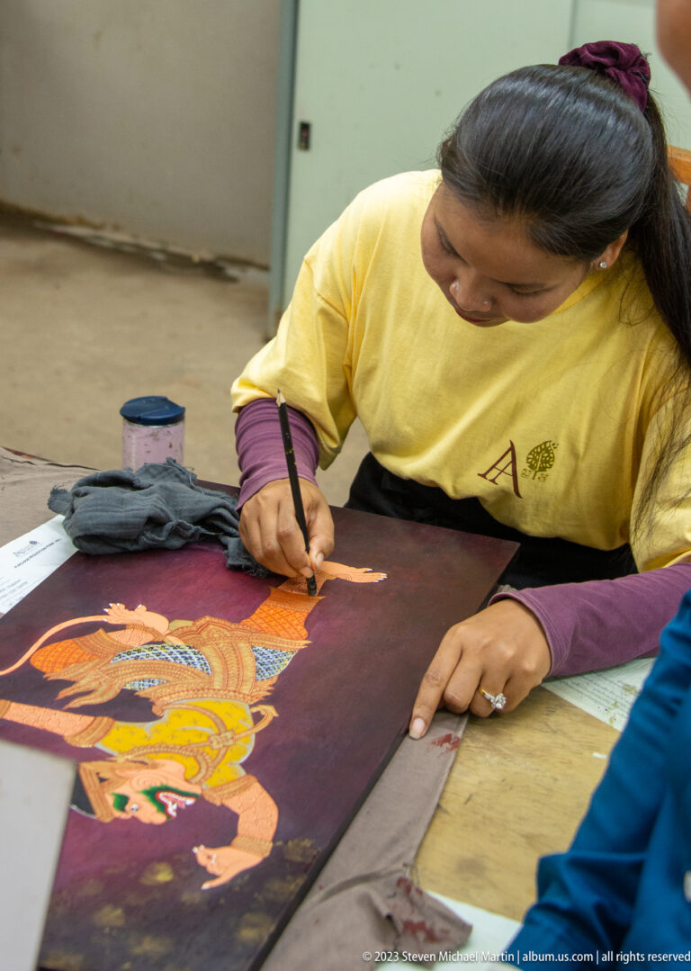 Woman using a fine brush to paint an orange Khmer-style figure on a dark maroon panel.