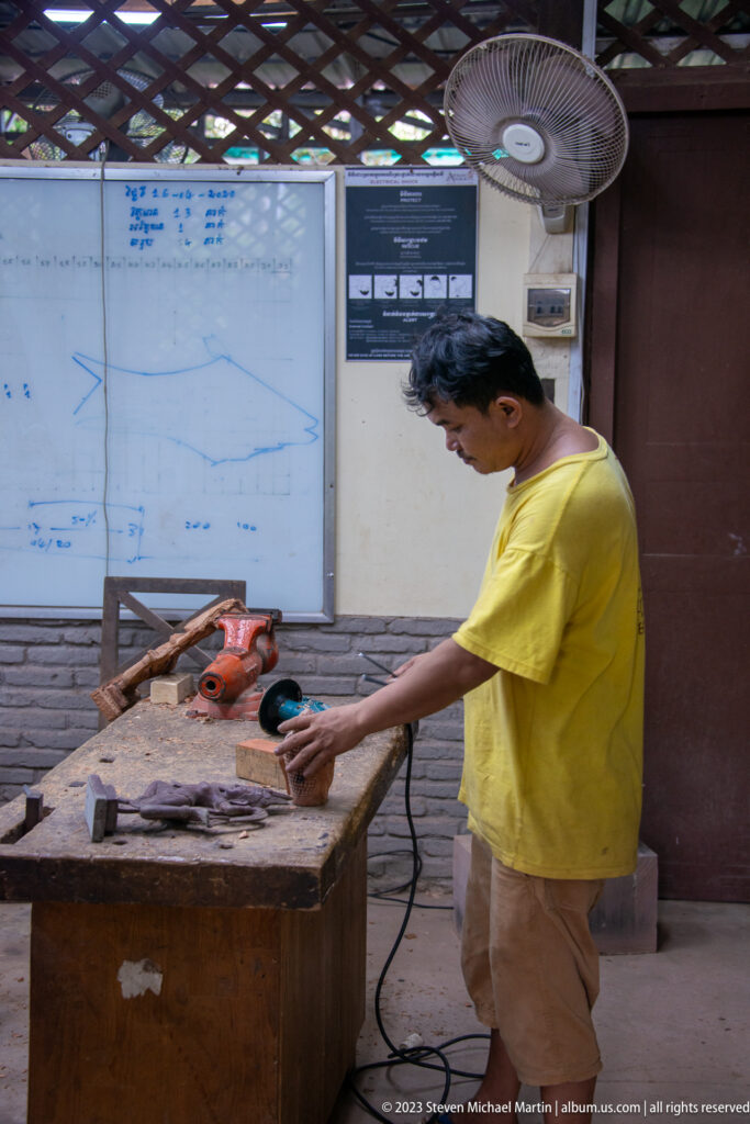 Man in a yellow shirt shaping a wooden block with an orange powered bench tool.