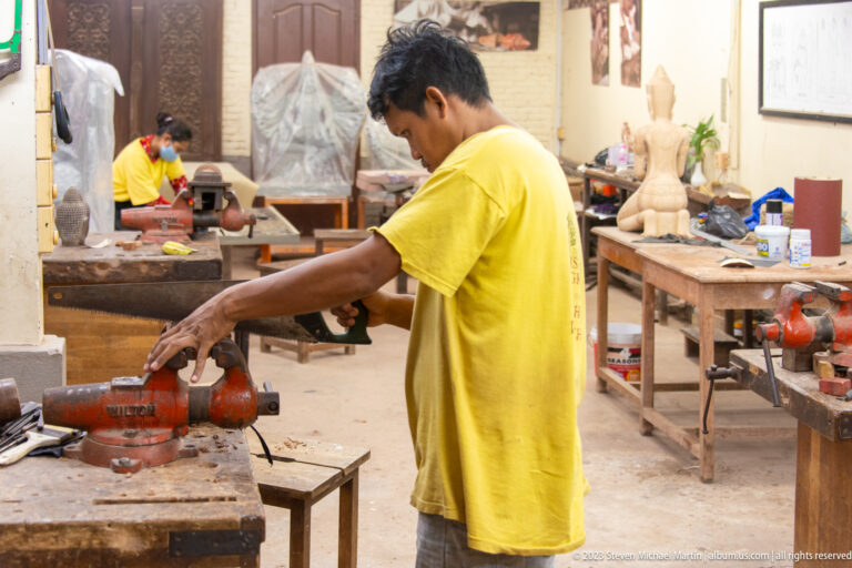 Man in a yellow shirt working at an orange wood machine in a busy carving workshop.