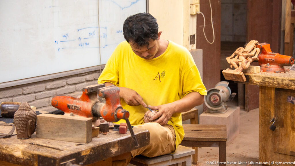 Seated artisan in a yellow shirt carving wood beside an orange machine and grinding wheel.