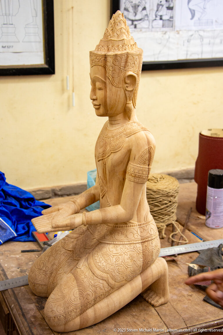 Unfinished wooden seated female figure with ornate jewelry, patterned garments, and extended hands.