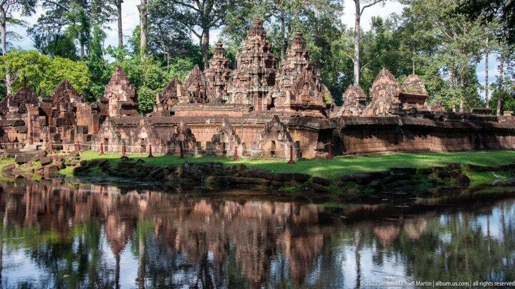 Oblique panorama of Banteay Srei's sanctuary towers and pediments reflected in water, with mossy stones along the bank.