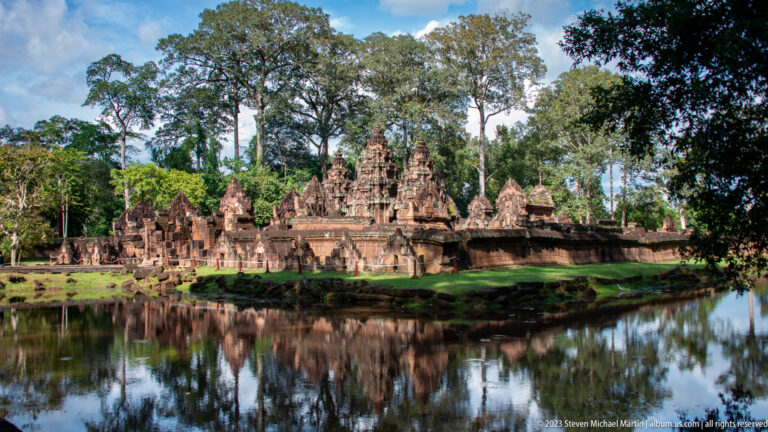 Wide landscape view of Banteay Srei's sanctuary towers, enclosure walls, water, and surrounding forest.