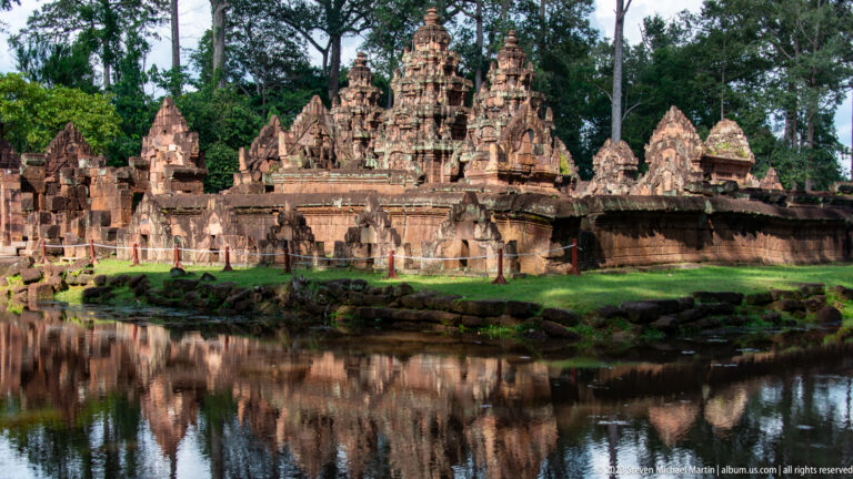 Closer three-quarter view of Banteay Srei's central sanctuary towers and carved pediments reflected in water.