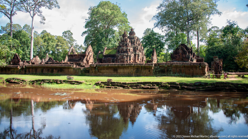 Wide view of Banteay Srei's towers and enclosure reflected in still water beneath surrounding trees.