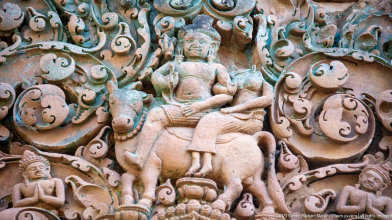 Close sandstone relief of an adorned male figure riding a hoofed animal amid curling foliage and smaller attendants.