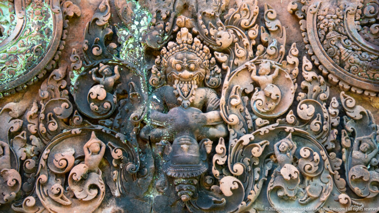 Fierce frontal guardian relief with bulging eyes, exposed teeth, raised arms, and curling sandstone foliage.