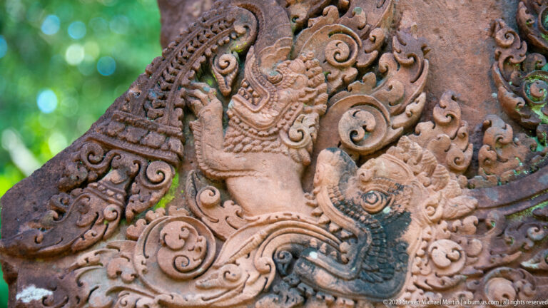Oblique close view of an adorned carved figure with a tall headdress amid curling sandstone ornament.