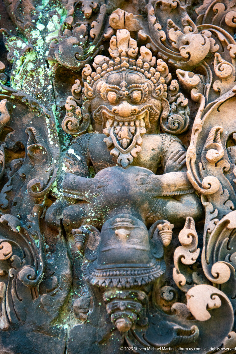 Extreme close-up of a fierce Khmer guardian face with round eyes, broad nose, exposed teeth, and floral ornament.