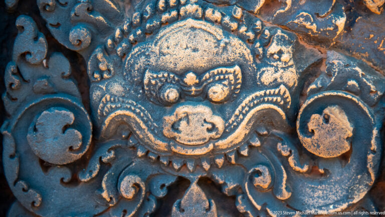 Close carved guardian face with round eyes, curved brows, small fangs, and symmetrical sandstone scrollwork.