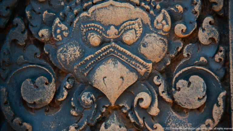 Stylized sandstone guardian mask with large round eyes, angular brows, pointed beaklike face, and symmetrical scrolls.