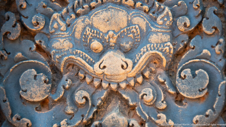 Close carved guardian face with bulging eyes, rounded snout, pointed fangs, and curling sandstone scrolls.