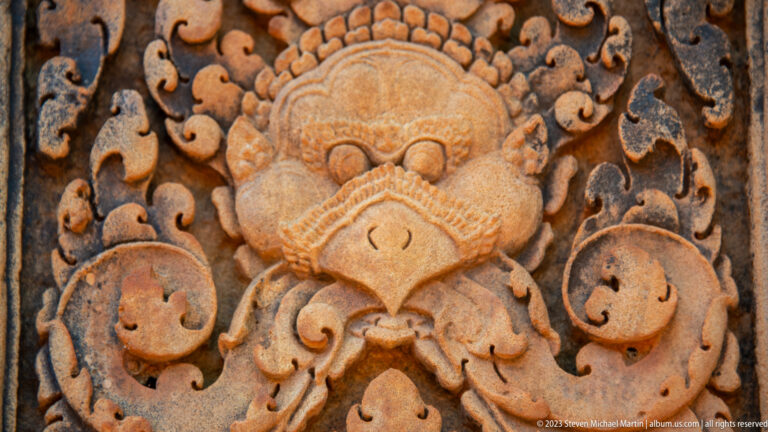 Weathered sandstone guardian mask with round eyes, pointed lower face, scalloped crown, and symmetrical scrolls.
