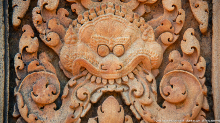 Close sandstone guardian mask with rounded eyes, broad snout, pointed teeth, and curling vegetal ornament.