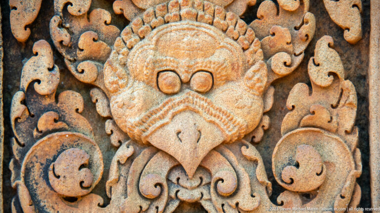 Close sandstone relief of a stylized guardian mask with round eyes, pointed beaklike face, and symmetrical scrollwork.