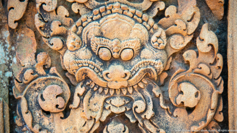 Close carved guardian face with bulging eyes, broad snout, pointed fangs, and curling sandstone foliage.