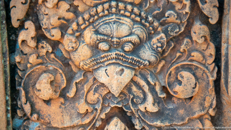 Dark-weathered sandstone guardian mask with round eyes, pointed lower face, and large surrounding scrolls.