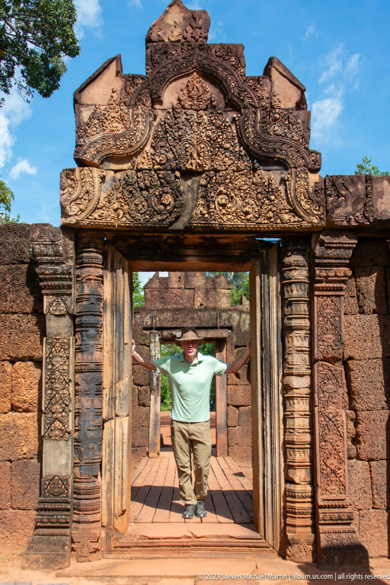 Visitor in a green shirt and brimmed hat standing with arms spread inside a carved temple doorway.