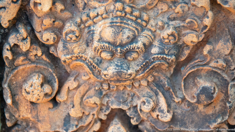 Weathered sandstone guardian face with bulging eyes, broad muzzle, exposed teeth, and curling ornament.