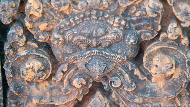 Dark-weathered sandstone guardian mask with round eyes, pointed lower face, scalloped crown, and broad side scrolls.