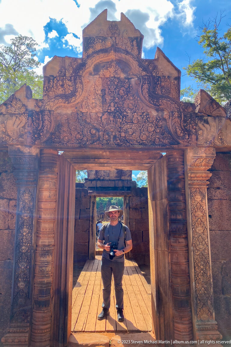 Visitor standing in a carved sandstone doorway on a wooden walkway at Banteay Srei.