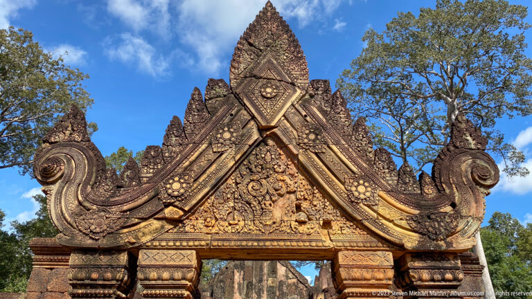 Low-angle view of an ornate Banteay Srei gateway with crossed pediment bands and curling stone volutes.