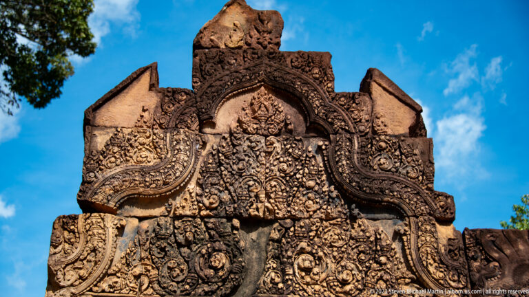 Upward close view of a richly carved Banteay Srei pediment against blue sky.