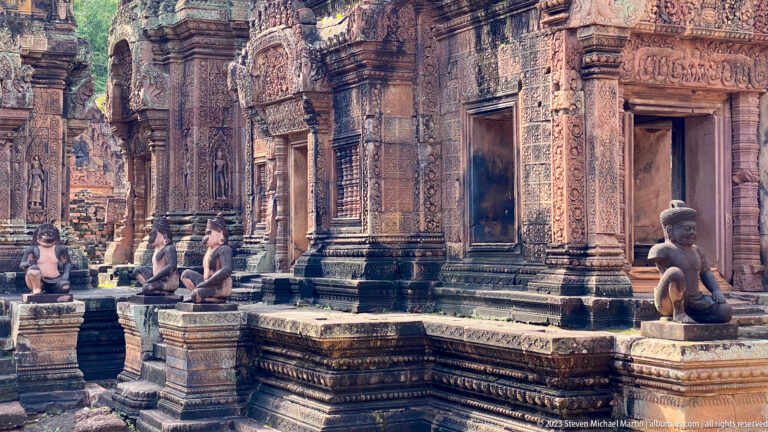 Seated stone guardians lining a raised platform before carved Banteay Srei sanctuary doorways.