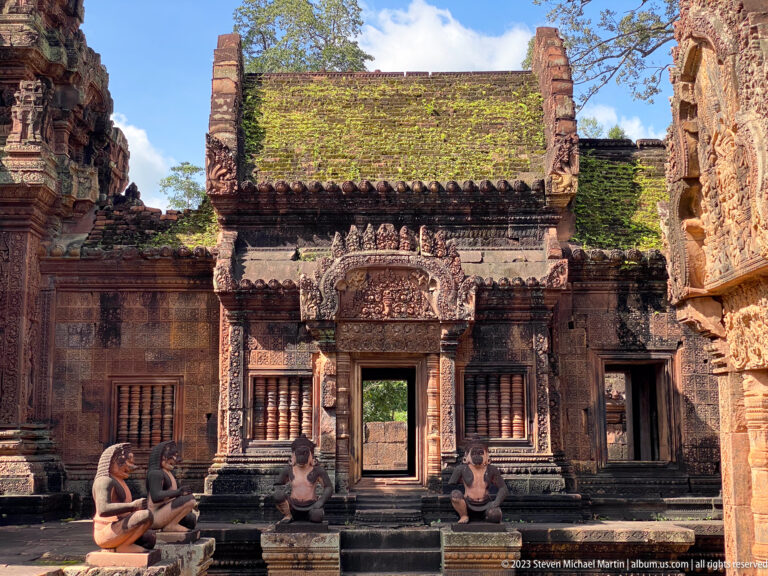Banteay Srei shrine façade with central doorway, stone-balustered windows, seated guardians, and mossy roof.