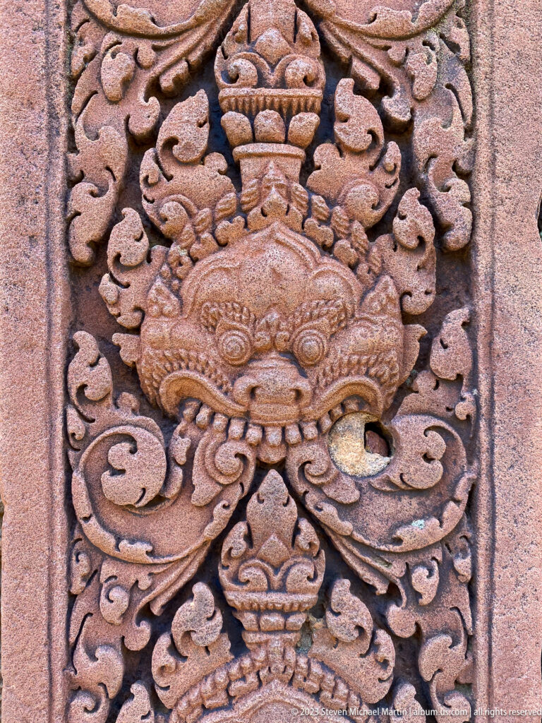 Close sandstone carving of a frontal kala monster mask surrounded by curling foliage.