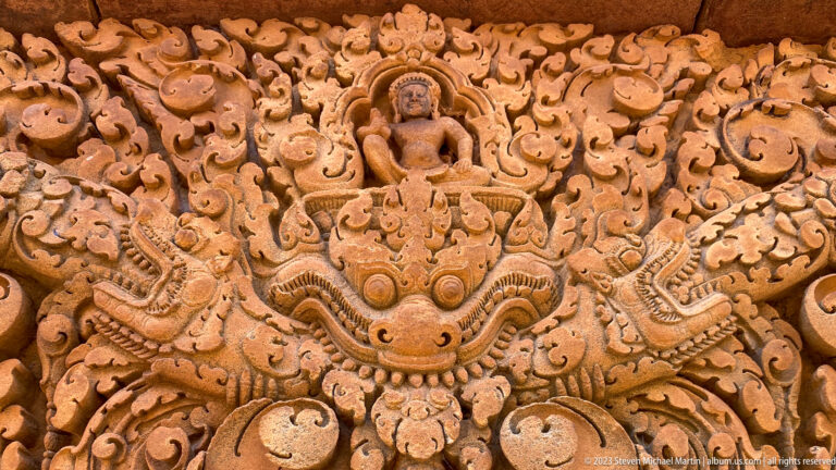 Symmetrical Banteay Srei lintel with seated deity, central kala mask, and paired mythical creatures.
